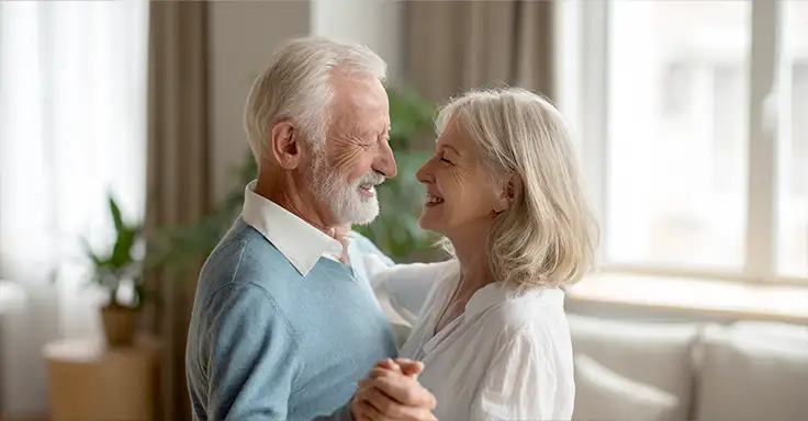 Älteres Paar tanzt lächelnd in einem sonnigen Wohnzimmer